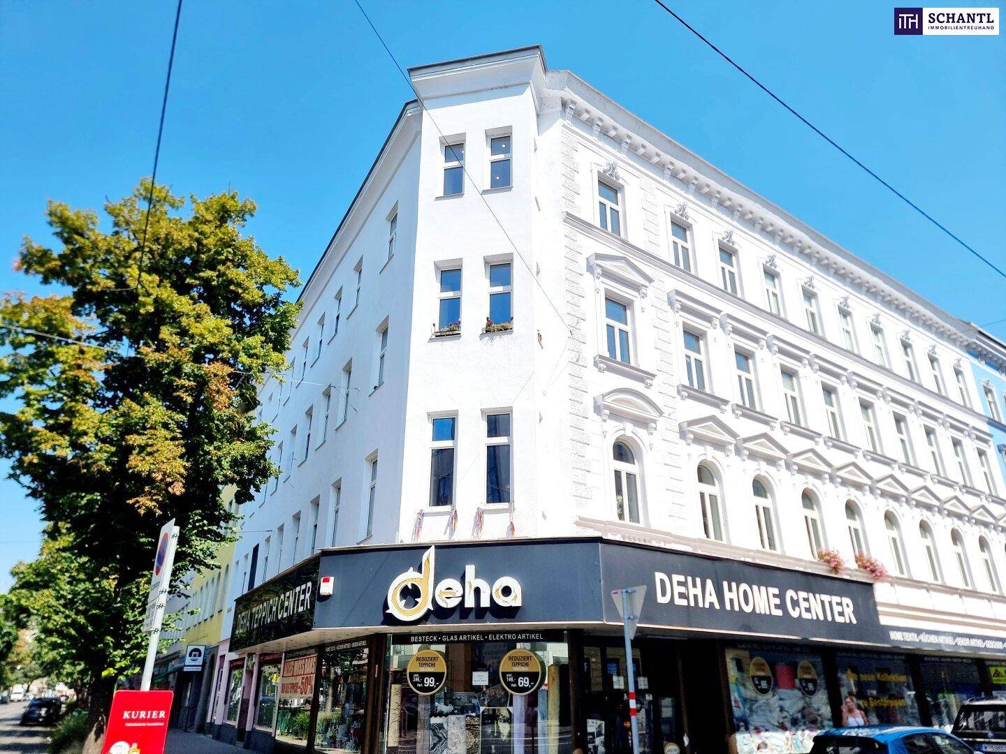 Eckgebäude mit weißer Fassade, Geschäften im Erdgeschoss und Bäumen an der Straße, sonniger Tag.
