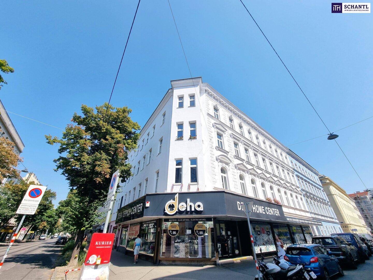 Eckgebäude mit weißer Fassade, Geschäften im Erdgeschoss und Bäumen an der Straße, sonniger Tag.
