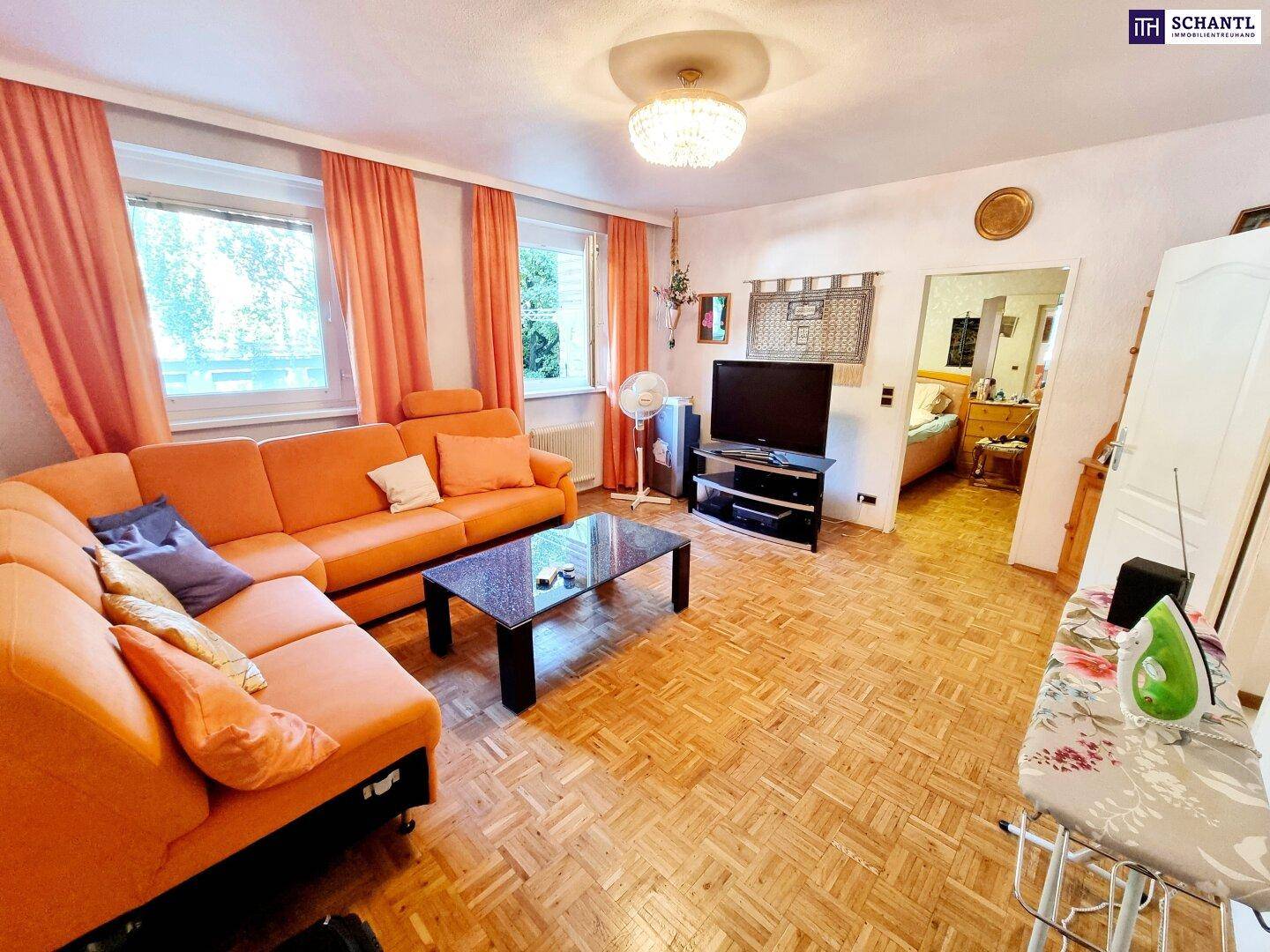 Helles Wohnzimmer mit orangefarbenem Ecksofa, Parkettboden und Blick ins Schlafzimmer.