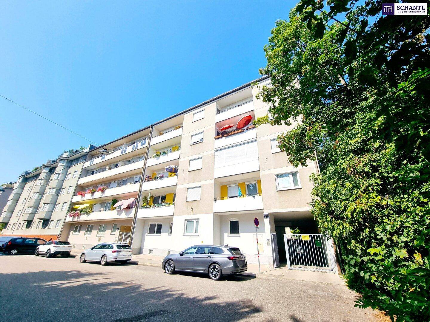 Mehrstöckiges Wohngebäude mit Balkonen und Parkplätzen an der Straße, umgeben von Grünflächen.