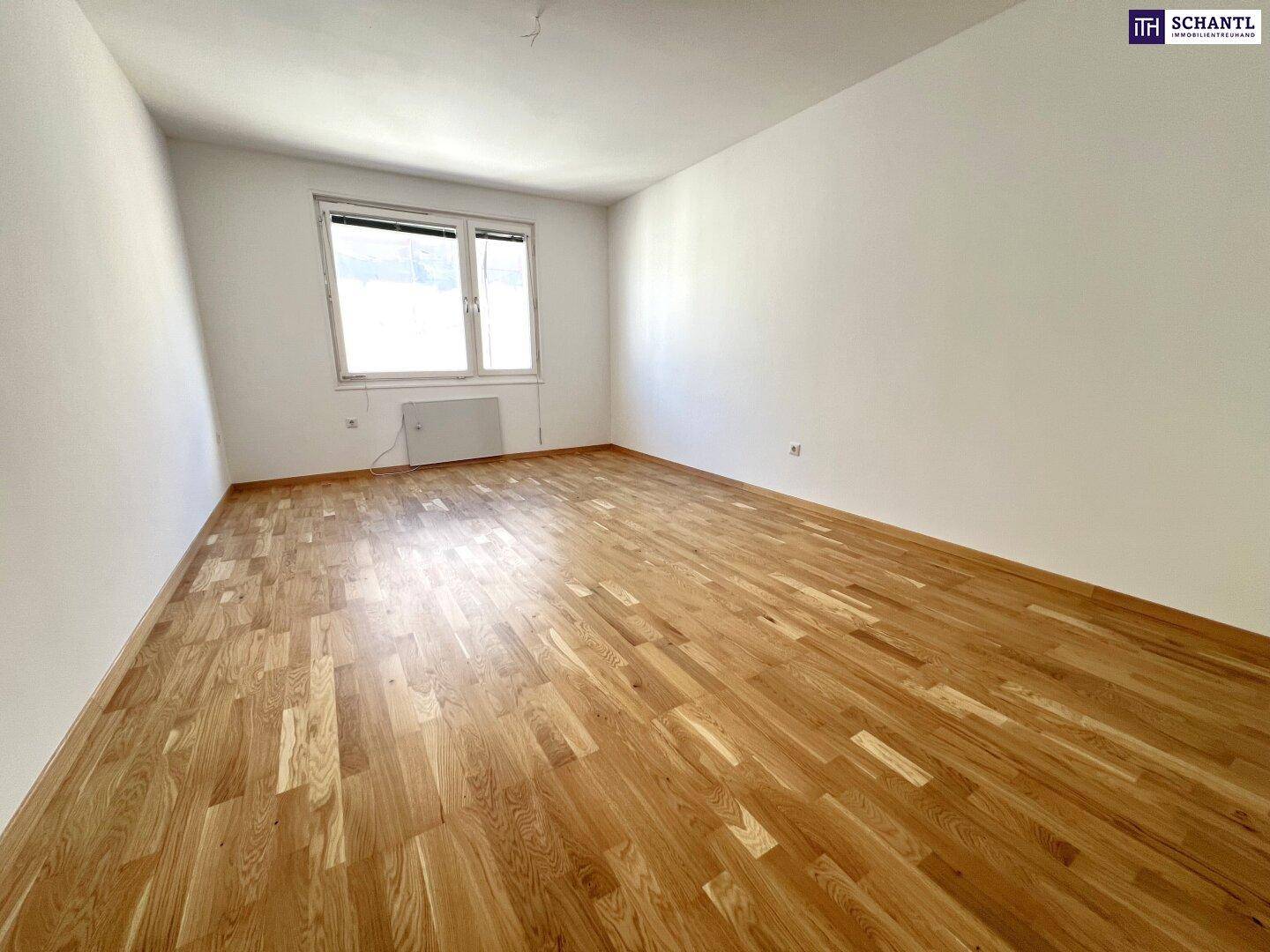 Leeres Zimmer mit Parkettboden und einem großen Fenster, das viel Tageslicht hereinlässt.