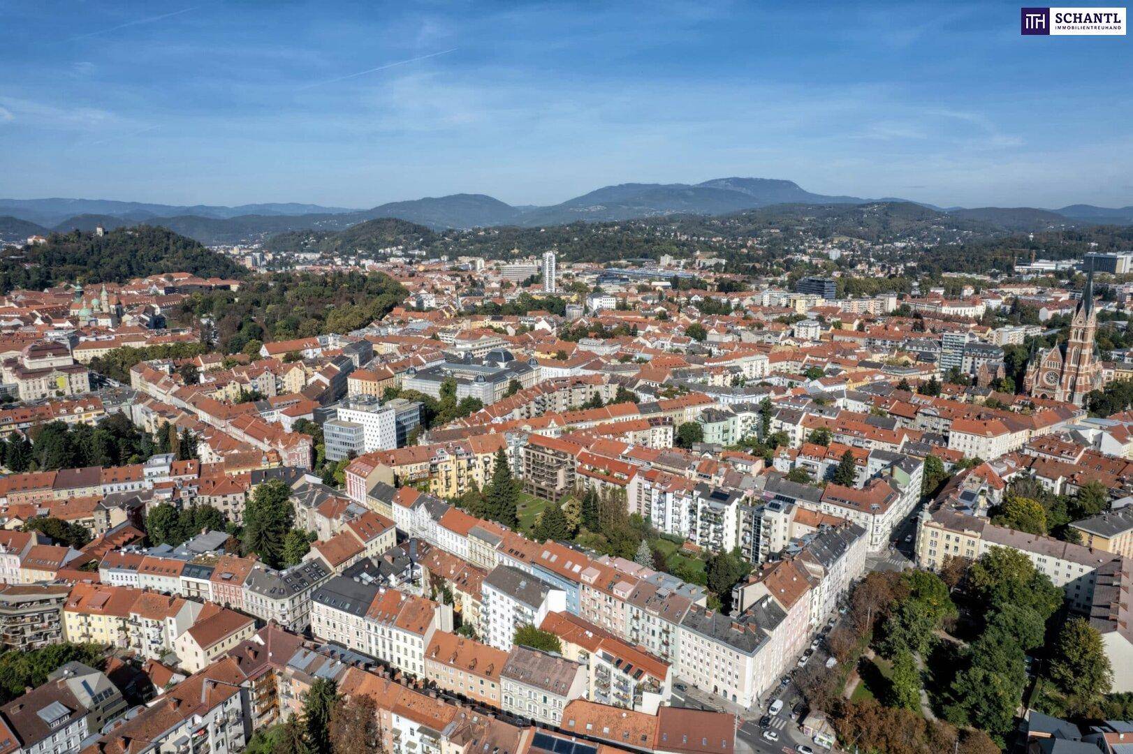 Weitläufiger Luftbildblick auf eine dicht bebaute Stadtlandschaft mit grünen Hügeln und Bergen im Hintergrund.