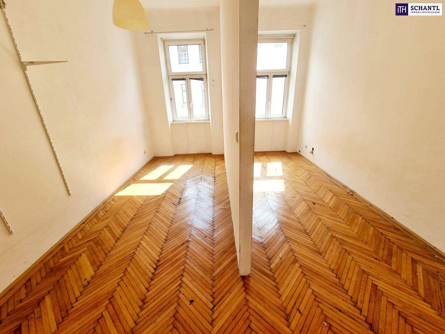 Leerer Raum mit Fischgrätparkett und zwei Fenstern, geteilt durch eine Trennwand.