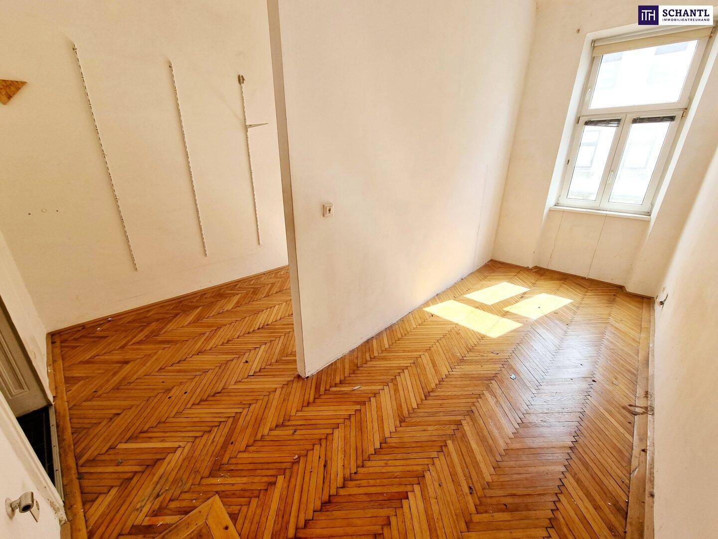 Heller Raum mit Fischgrätparkett und einem Fenster, teilweise durch eine Wand getrennt.