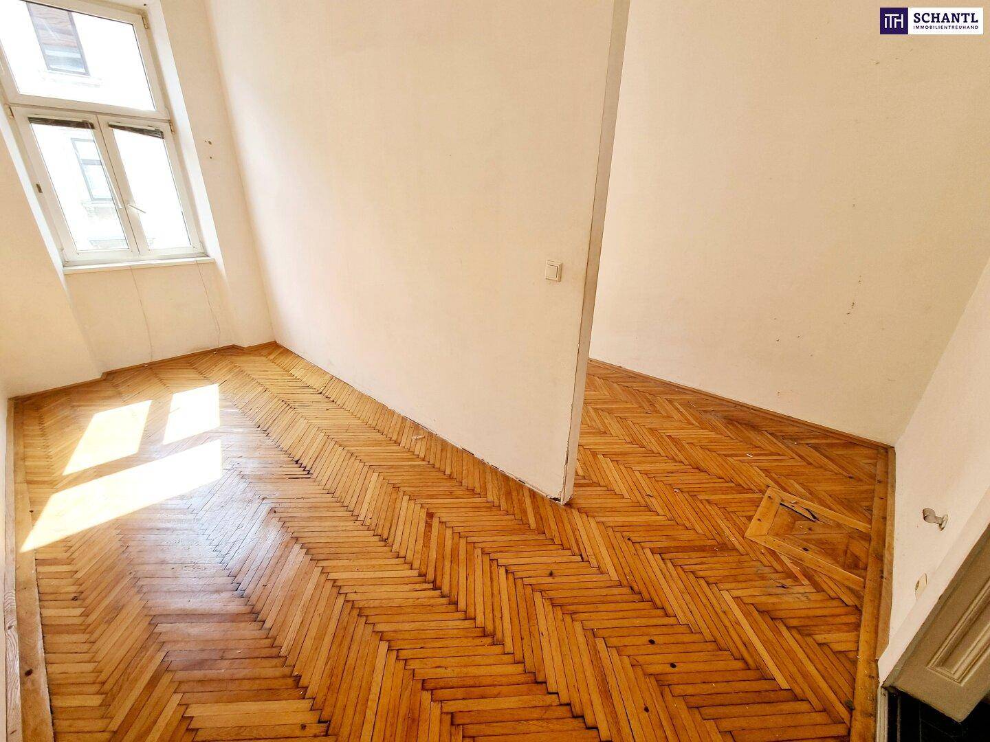 Leerer Raum mit schrägem Fenster und Fischgrätparkett, durch eine Wand geteilt.