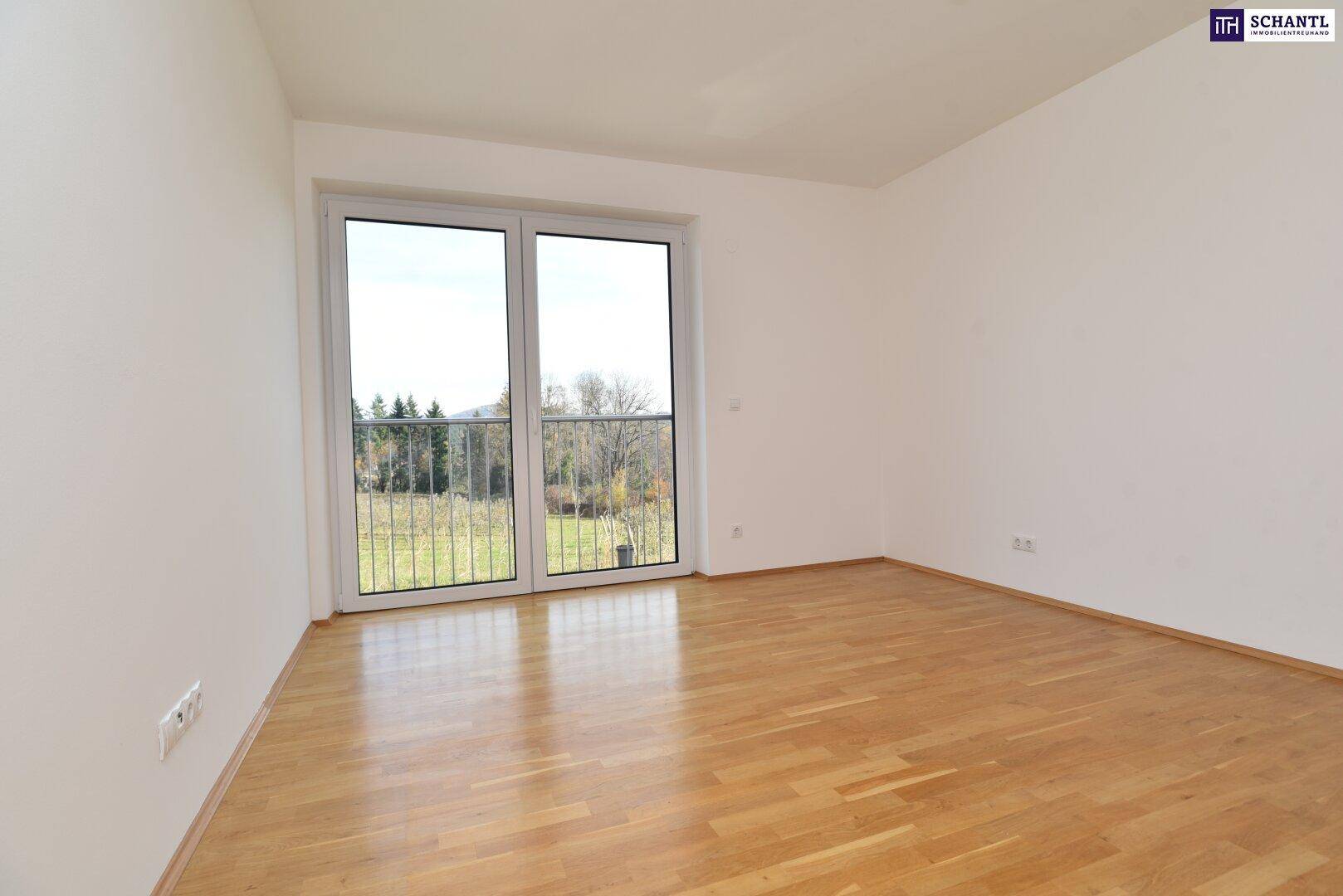Leeres Zimmer mit Parkettboden und großem Fenster mit Blick ins Grüne und Balkonzugang.