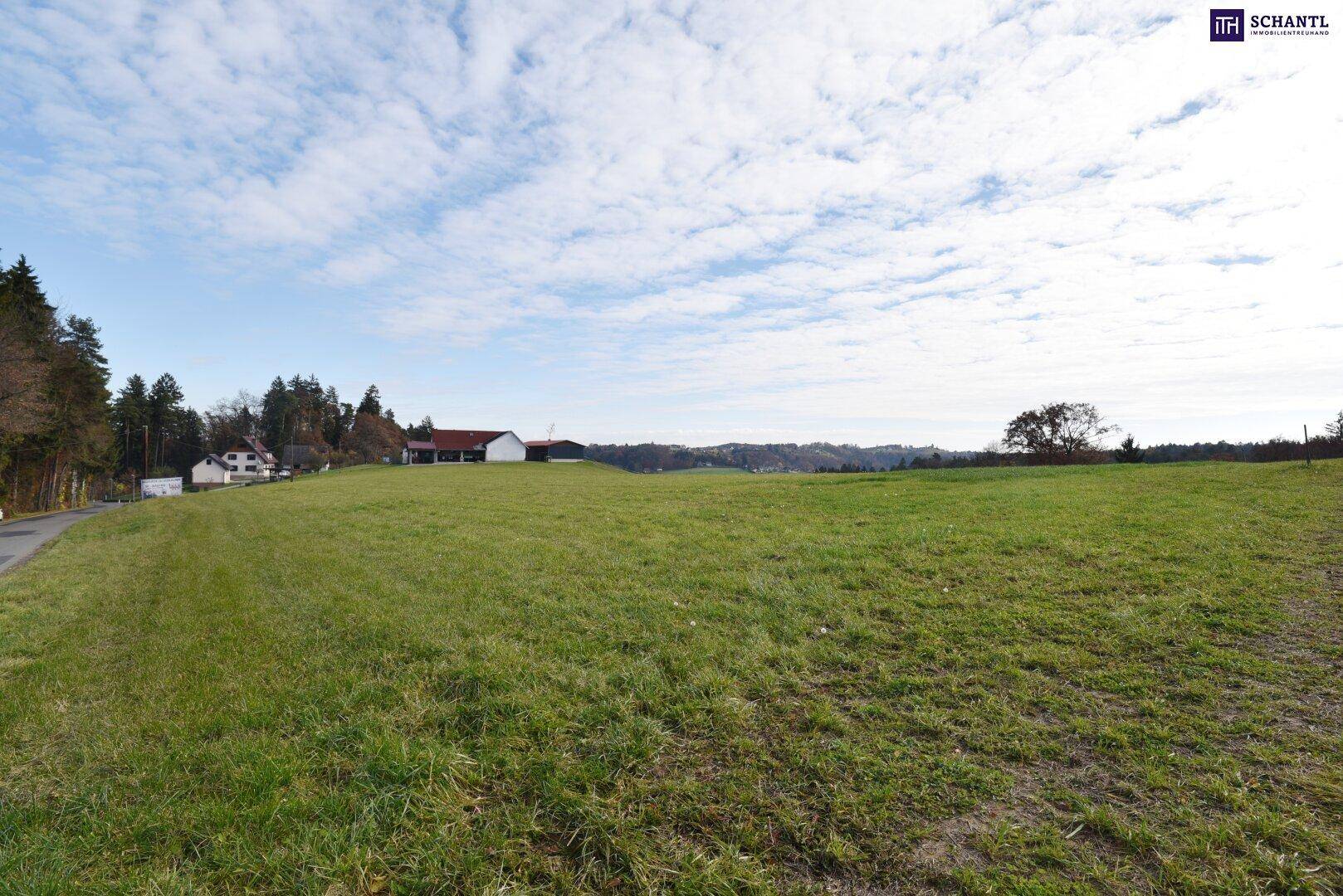 Weitläufiges grünes Feld mit Blick auf entfernte Gebäude und eine bewaldete Hügellandschaft.