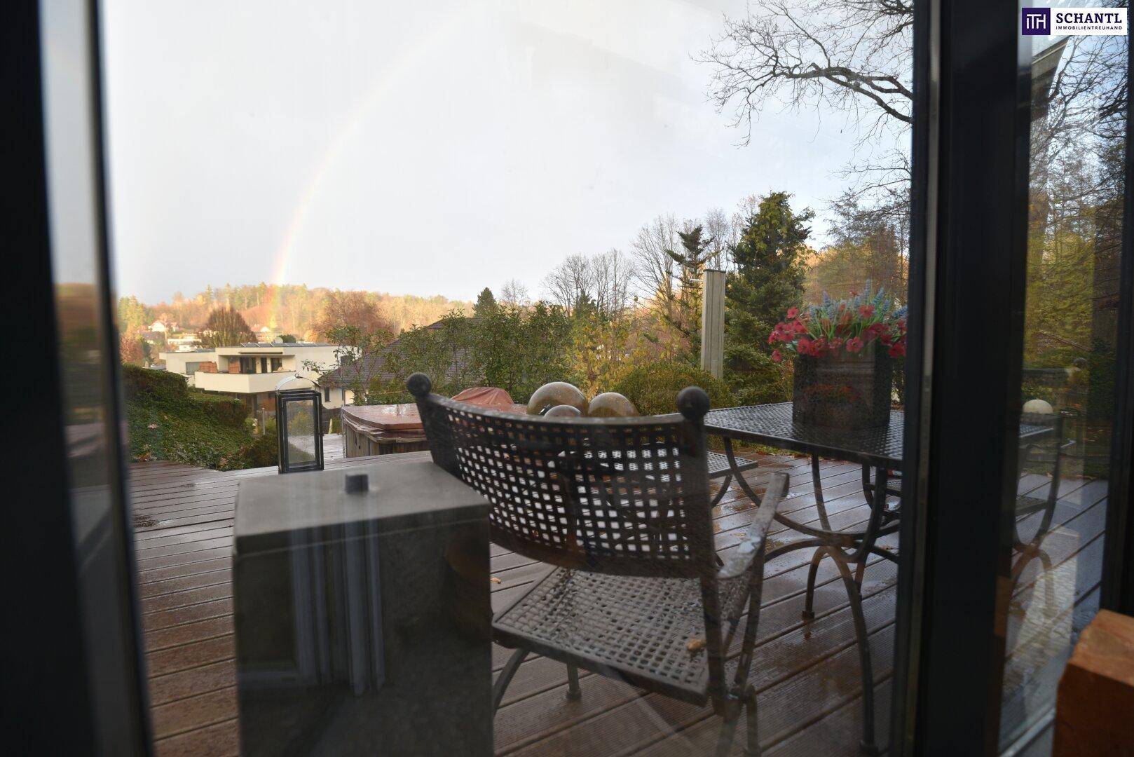 Blick von innen auf eine Holzterrasse mit Gartenmöbeln und einem Regenbogen am Horizont.