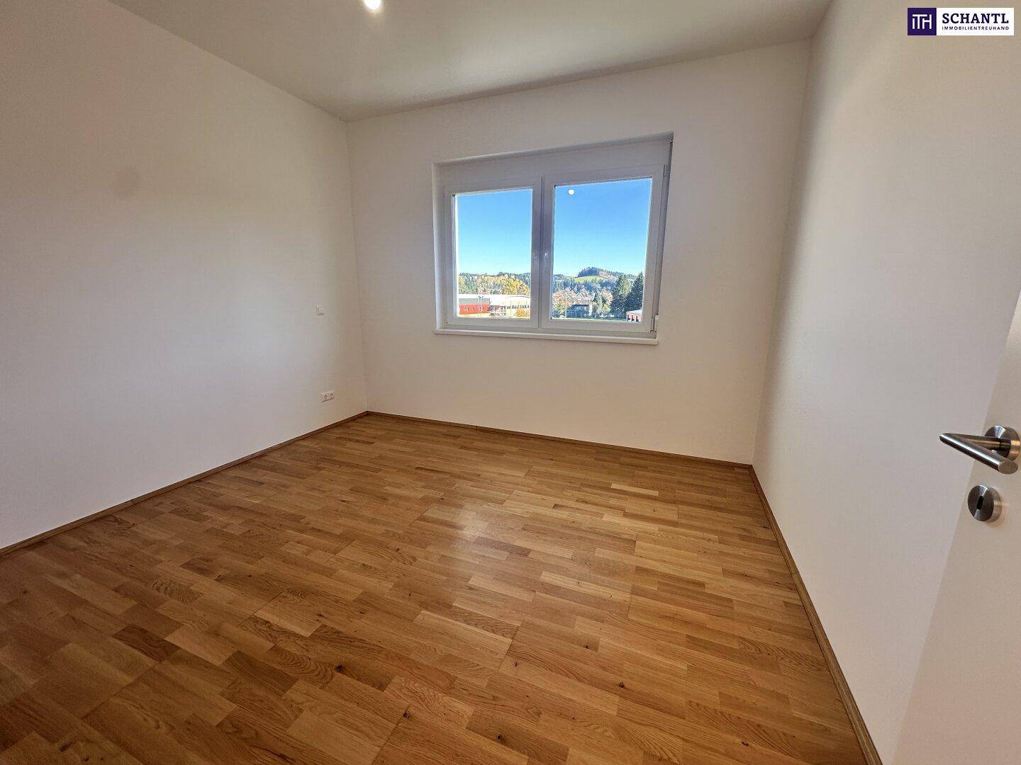 Leerer Raum mit hellem Parkettboden und Fenster, das einen schönen Ausblick in die Natur bietet.
