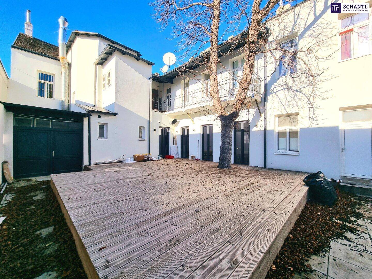 Sonniger Innenhof mit Holzterrasse und einem Baum, umgeben von Gebäudefassaden.