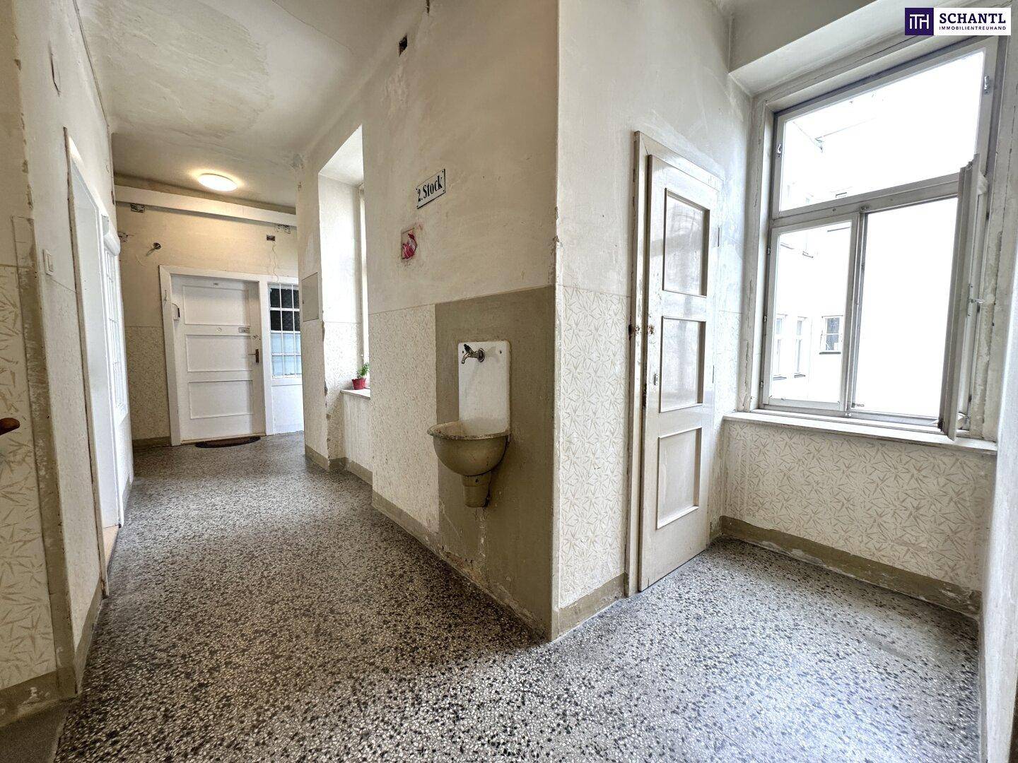Langer Flur mit Terrazzoboden, hohen Decken und einem alten Waschbecken.