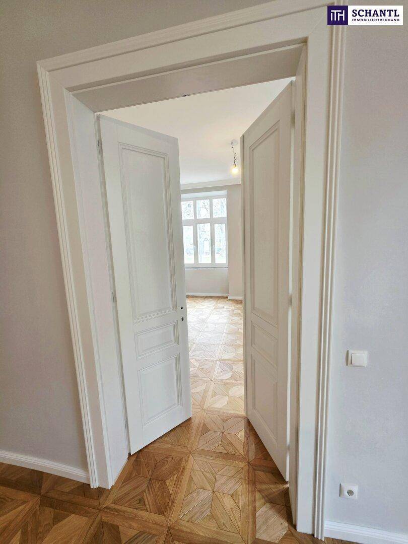 Weiße Doppeltüren führen in ein helles Zimmer mit Parkettboden und hohen Decken.