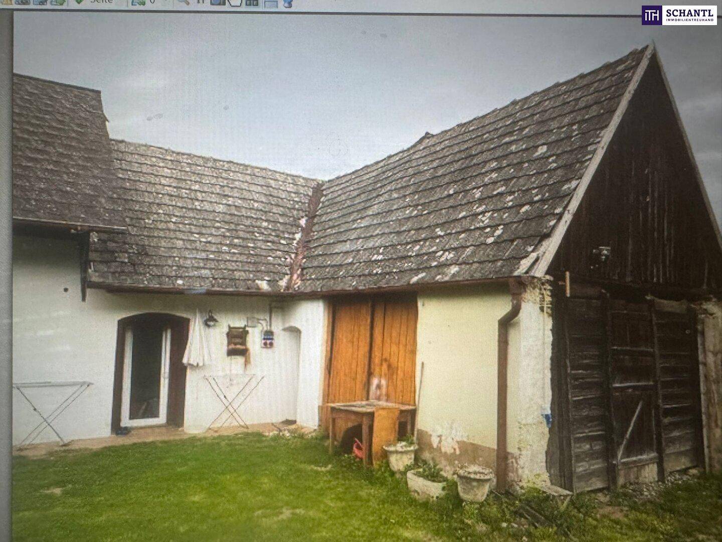 Rückseite des Gebäudes mit altem Dach, Holztüren und einem kleinen Gartenbereich, der viel Potenzial bietet.