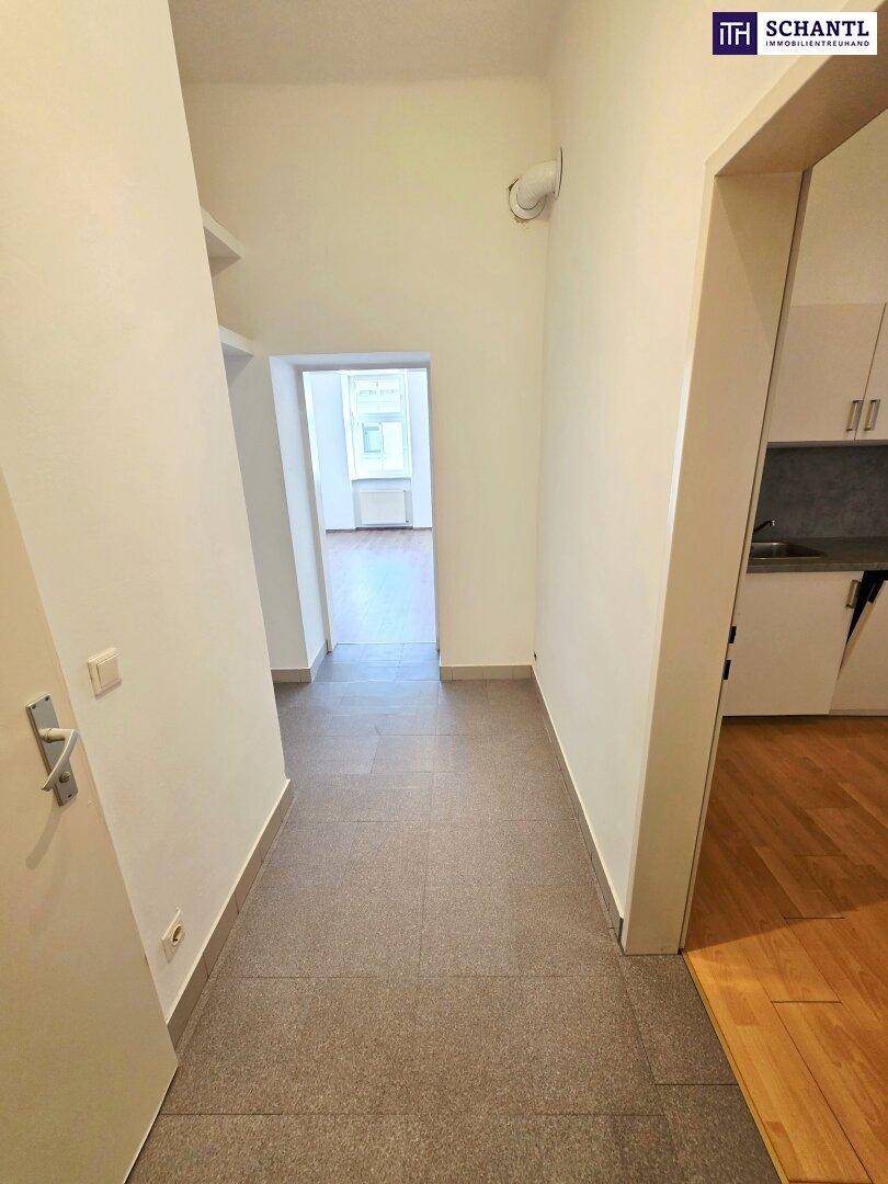 Heller Flur mit grauen Fliesen, der zu einem weiteren Zimmer mit Holzboden führt.