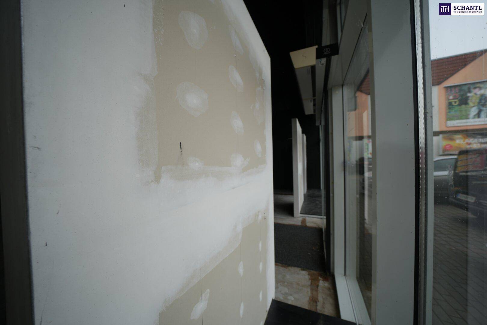 Blick von innen auf die großen Fensterfronten des Eingangs, mit einer teilweise verkleideten Wand im Vordergrund.
