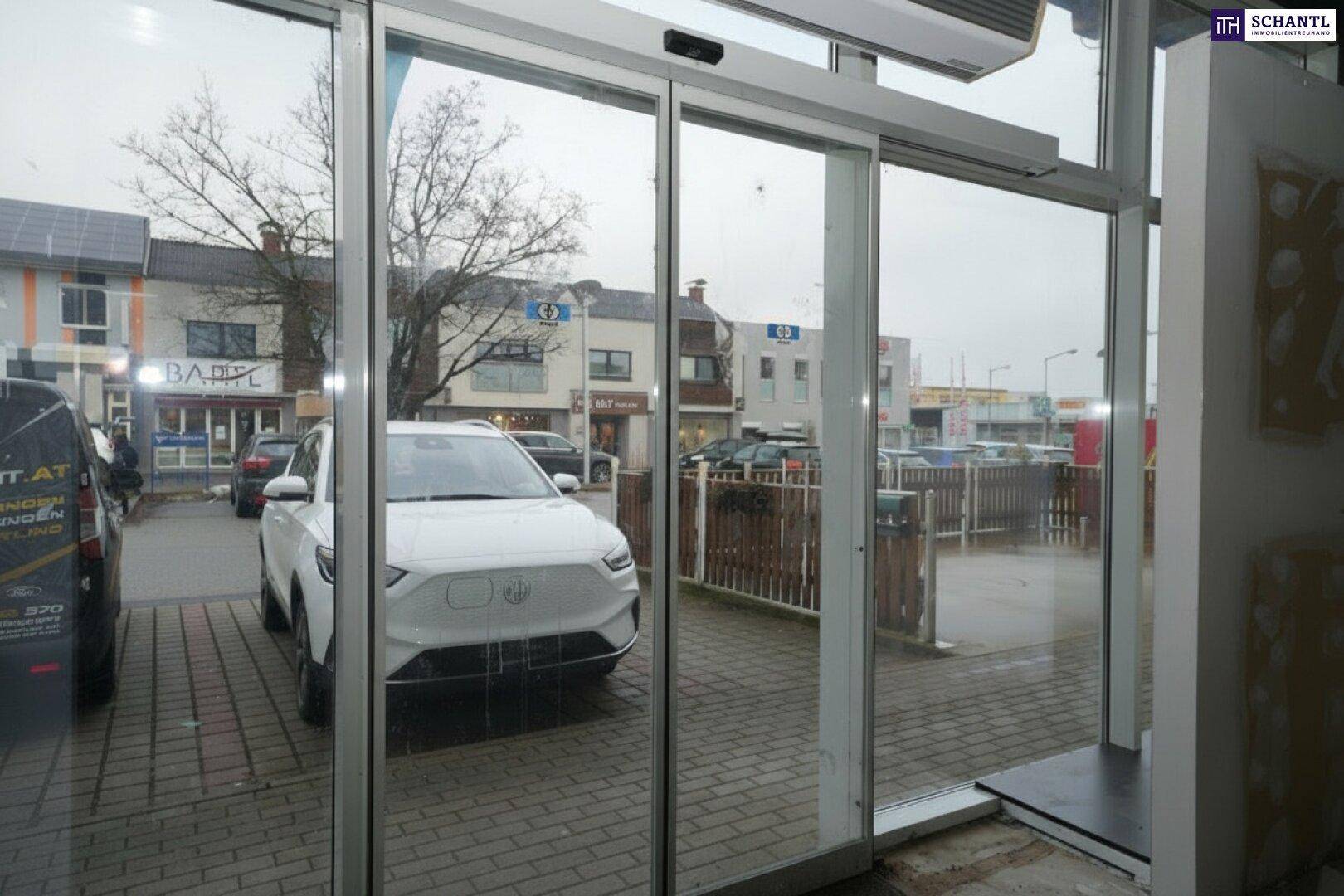 Blick aus dem Gewerberaum durch die automatischen Glastüren auf die belebte Straße und parkende Autos.
