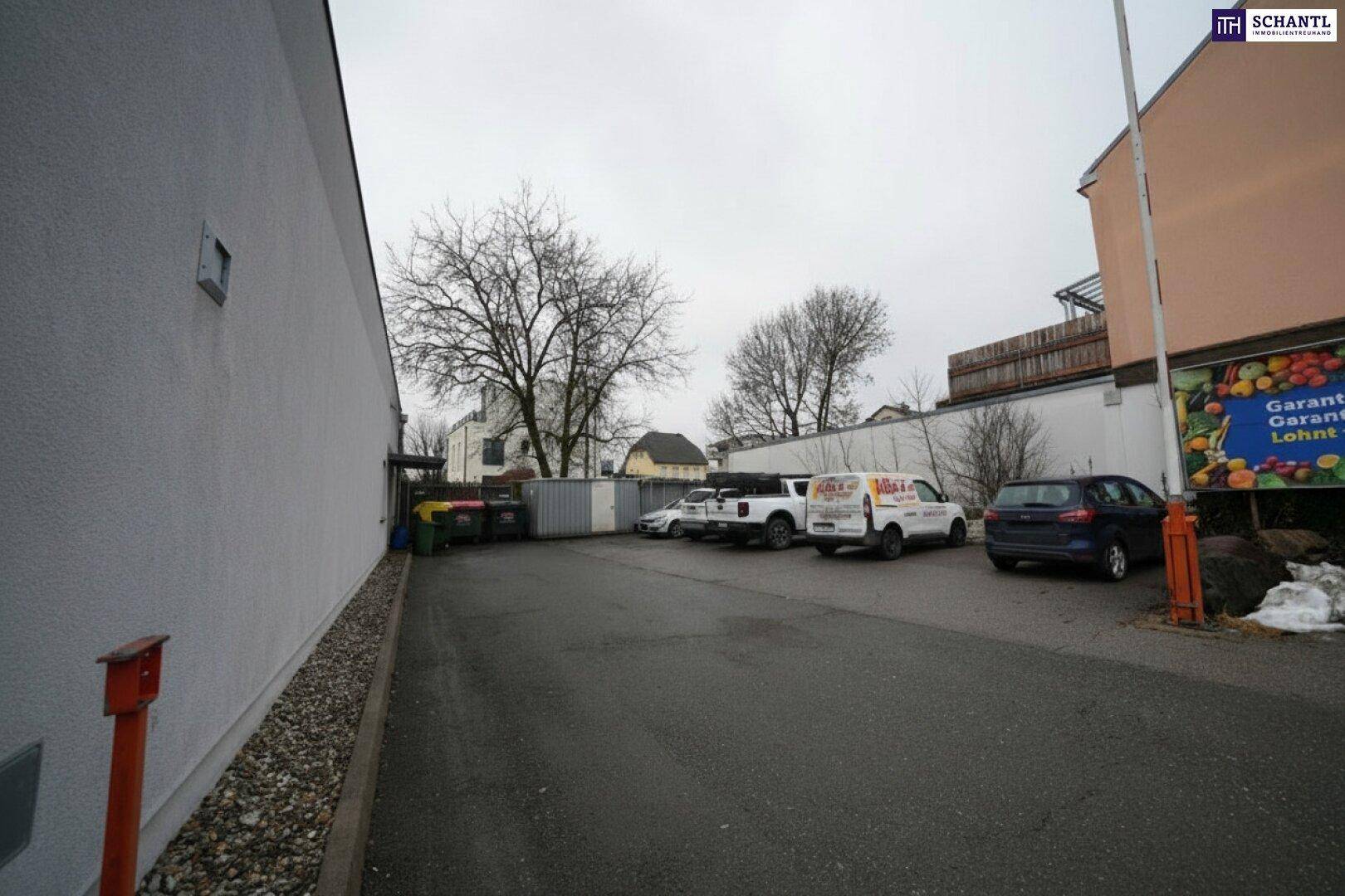 Außenbereich mit Parkplätzen und Mülltonnen, neben dem Gebäude gelegen, unter bewölktem Himmel.