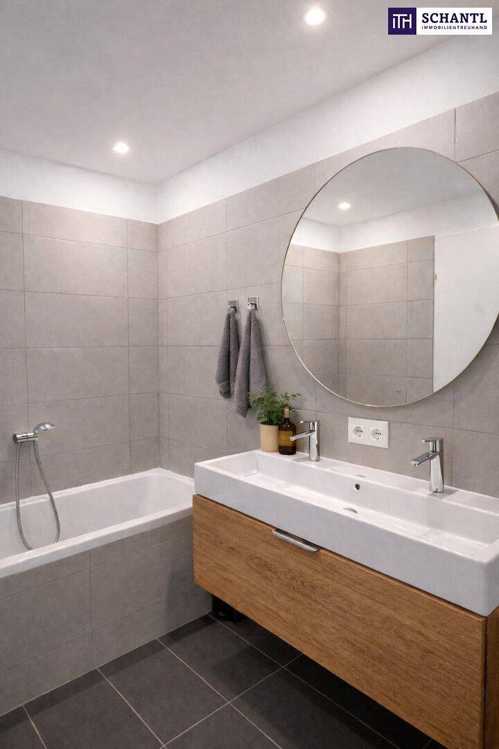 Modernes Badezimmer mit grauen Fliesen, Badewanne, Doppelwaschbecken und rundem Spiegel.