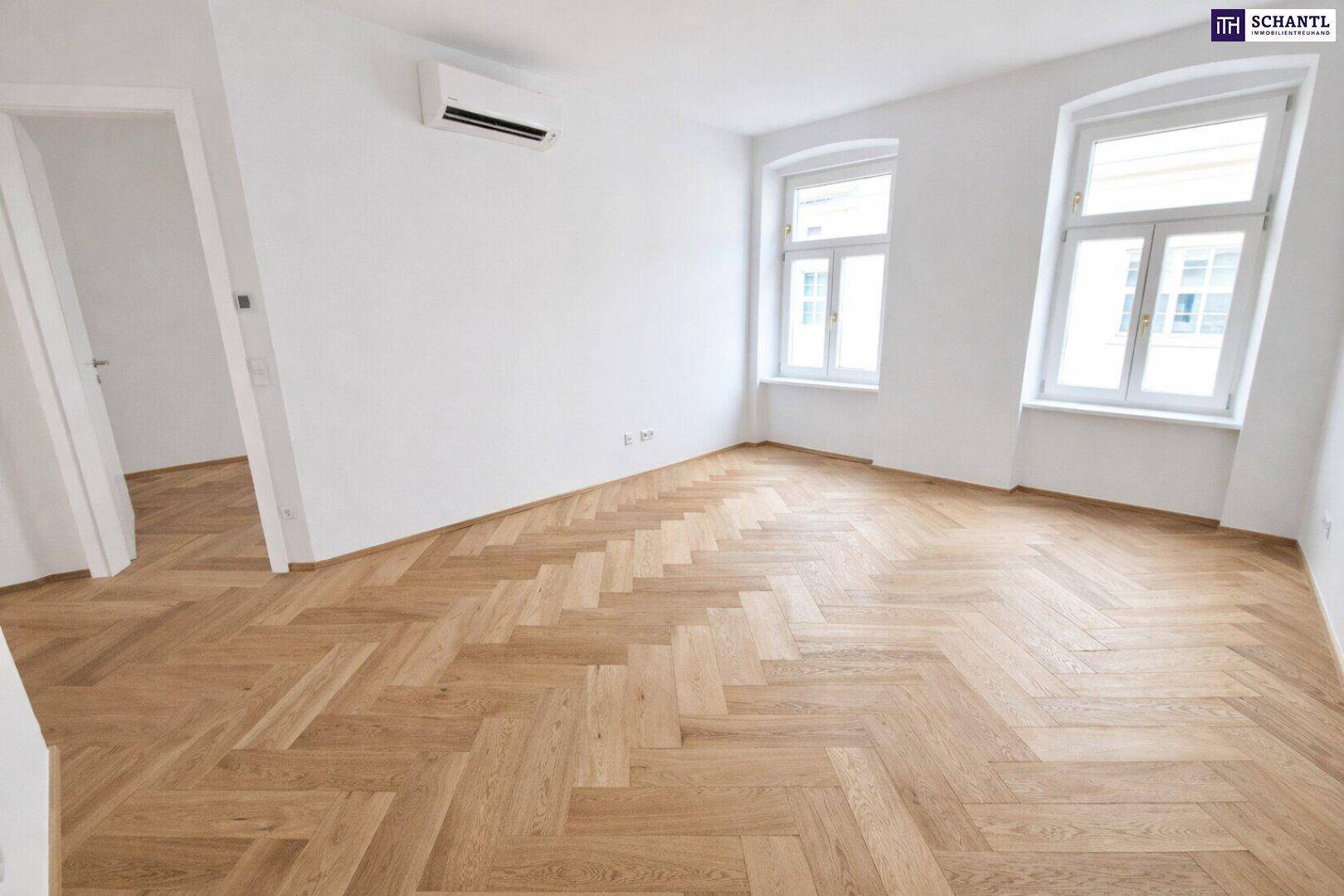 Geräumiger, heller Raum mit Fischgrätparkett, Klimaanlage und zwei großen Fenstern.