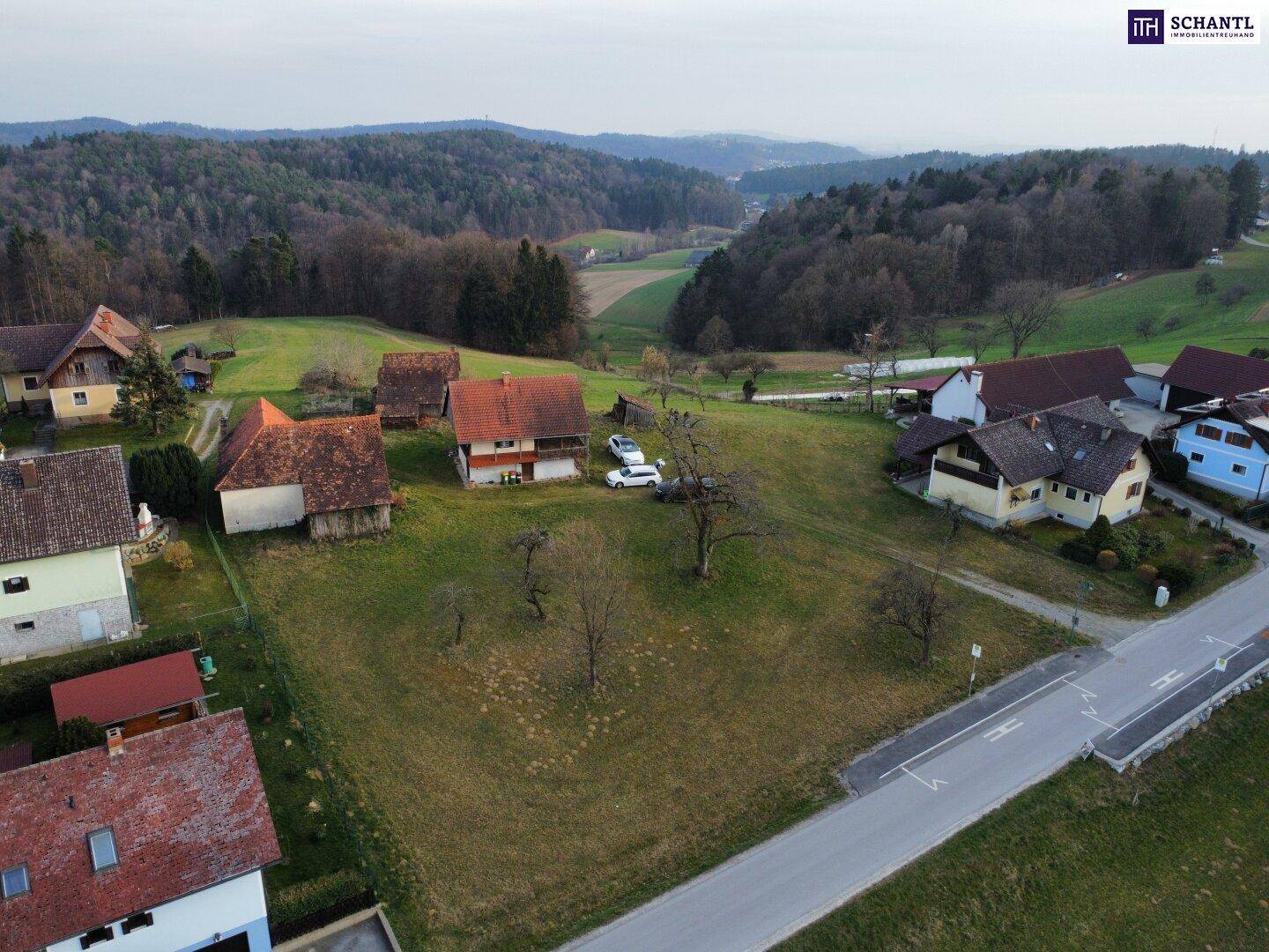 Weitläufige Luftaufnahme eines Wohngebiets mit Feldern, Häusern und bewaldeten Hügeln in der Ferne.