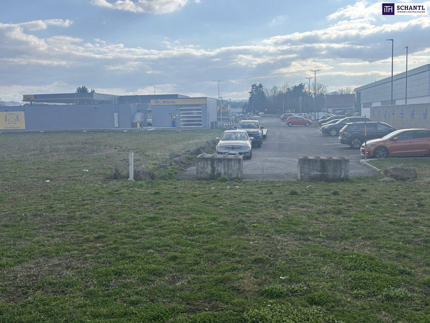 Blick auf ein Gewerbegebiet mit einer Autowaschanlage, Parkplätzen und geparkten Fahrzeugen unter bewölktem Himmel.