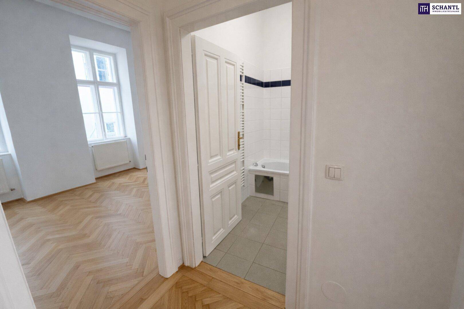 Blick vom Flur mit Fischgrätparkett in ein Badezimmer mit Badewanne und Fliesenboden.