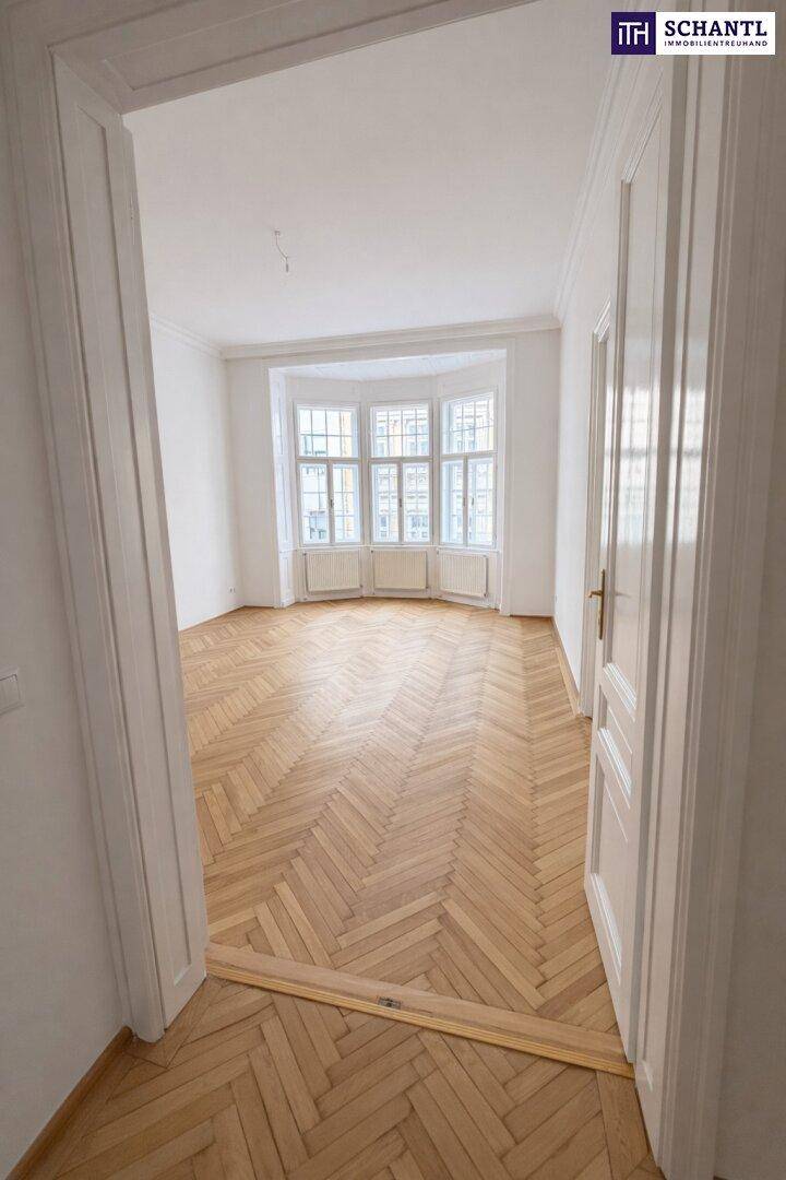 Blick vom Flur mit Fischgrätparkett in ein geräumiges Zimmer mit Erkerfenstern.