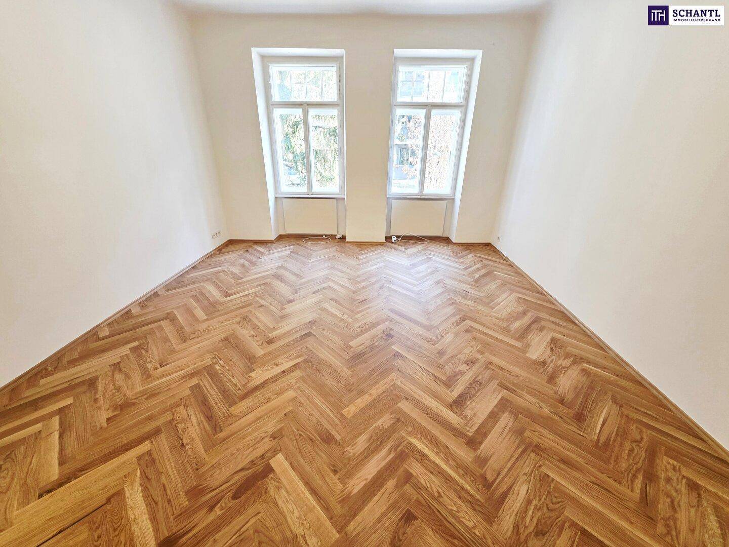 Großzügiges Zimmer mit Fischgrätparkett und zwei hohen Fenstern für viel Tageslicht.