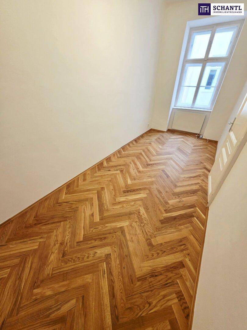 Helles Zimmer mit Fischgrätparkett und einem hohen Fenster, das den Raum erhellt.