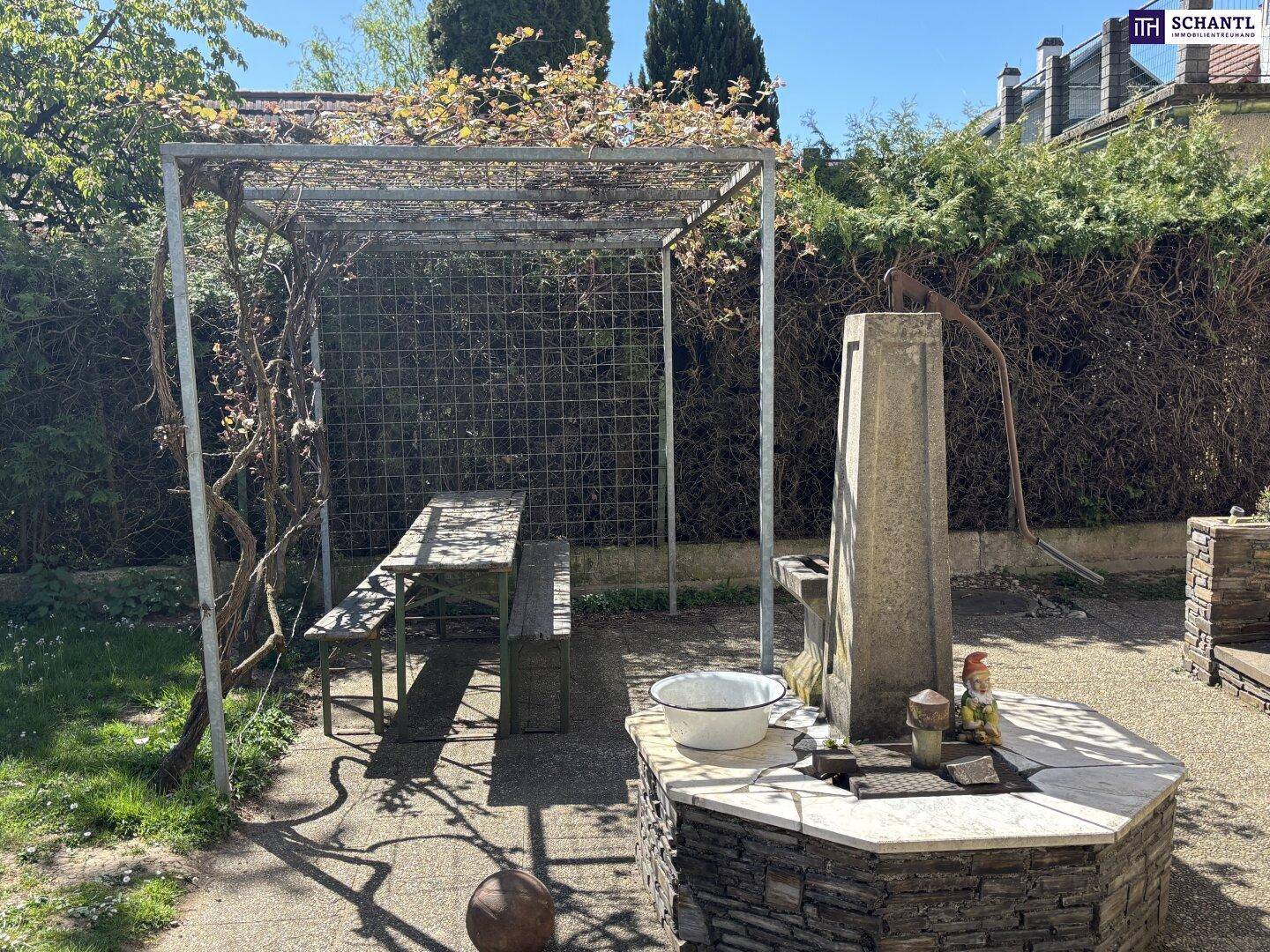 Gemütlicher Gartenbereich mit Pergola, Holztisch und einem dekorativen Brunnen.