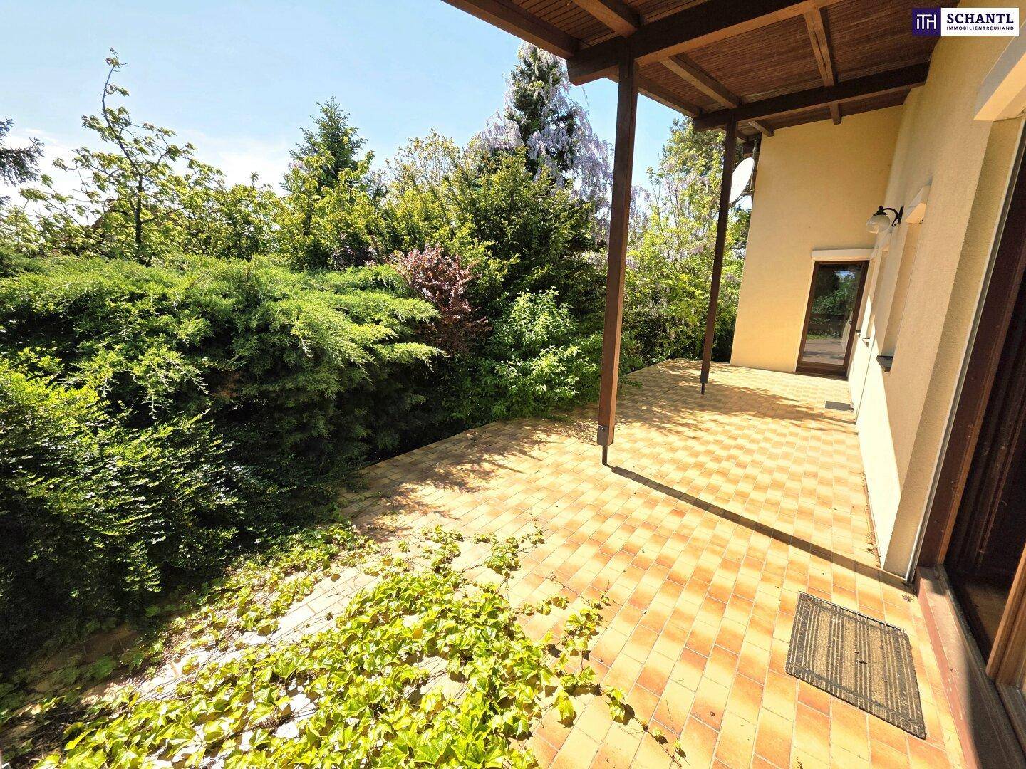 Große überdachte Terrasse mit Fliesenboden und üppiger Bepflanzung im Gartenbereich.