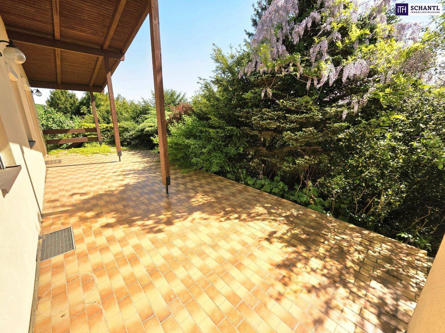 Sonnige, überdachte Terrasse mit Fliesenboden und umgebender grüner Gartenlandschaft.