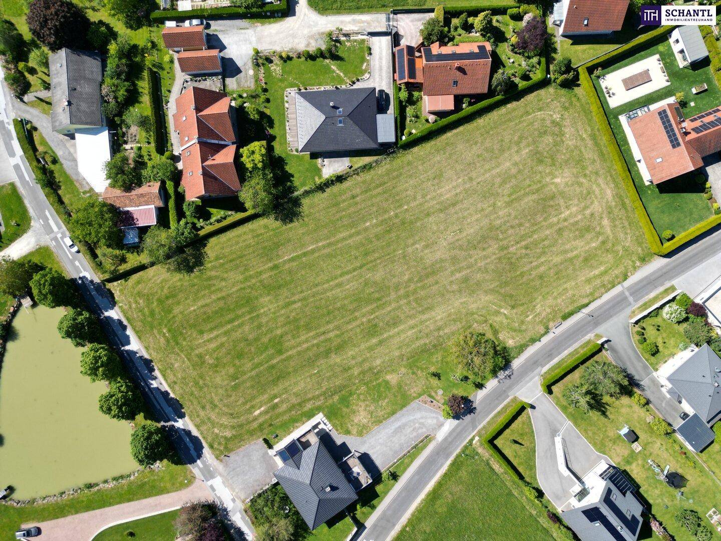 Luftaufnahme eines großen unbebauten Grundstücks, umgeben von Wohnhäusern, Straßen und einem Teich in ländlicher Umgebung.