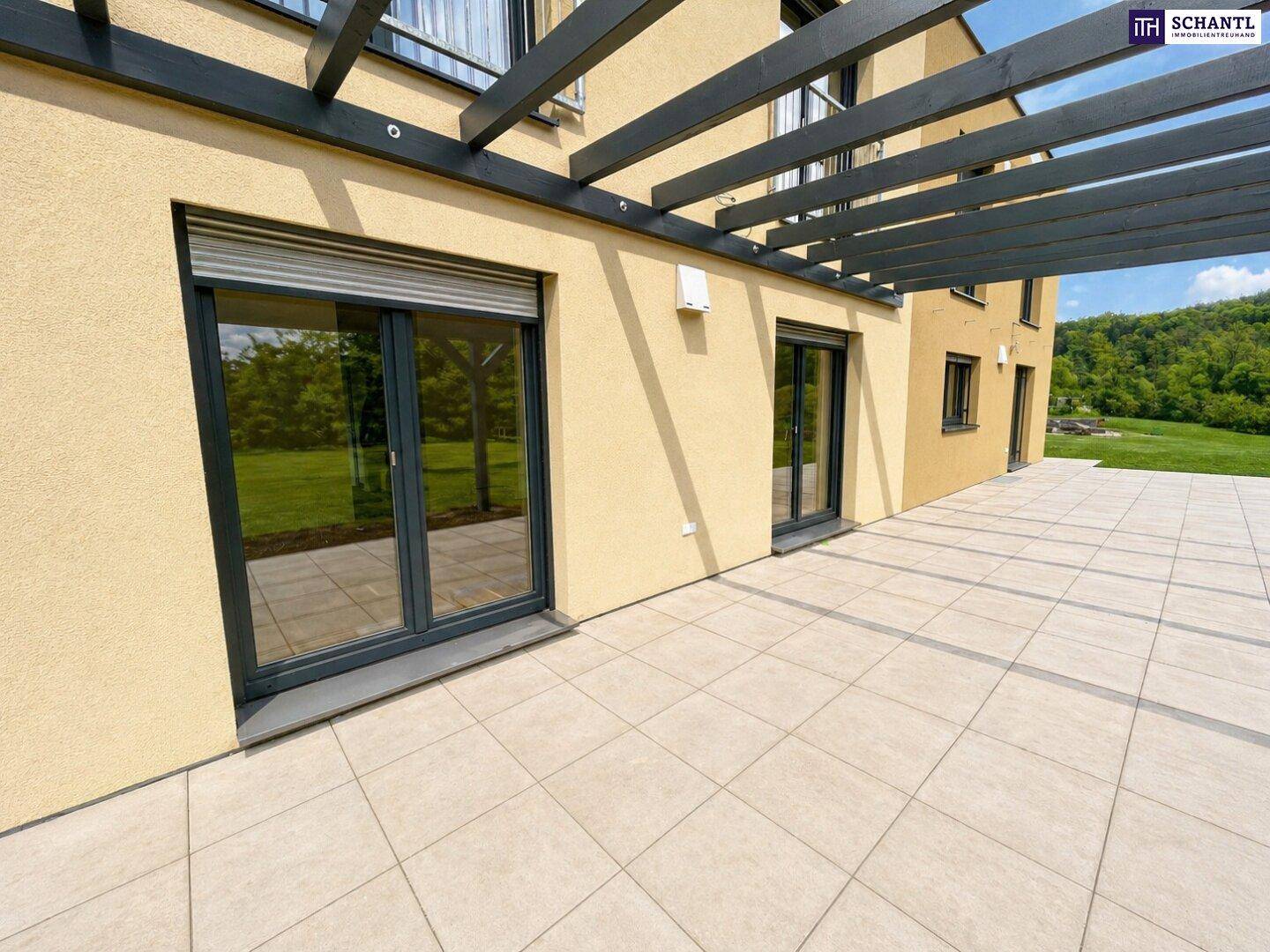 Große geflieste Terrasse mit Holzpergola und Zugang zum Haus, umgeben von grüner Landschaft.