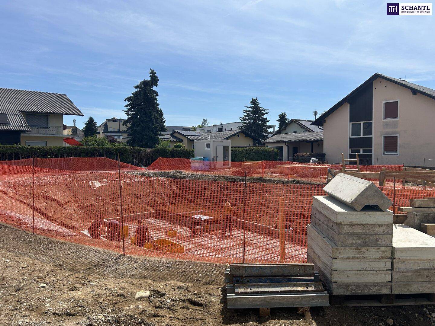 Baustelle mit vorbereitetem Fundament, Bewehrung und gestapelten Betonfertigteilen unter strahlendem Himmel.