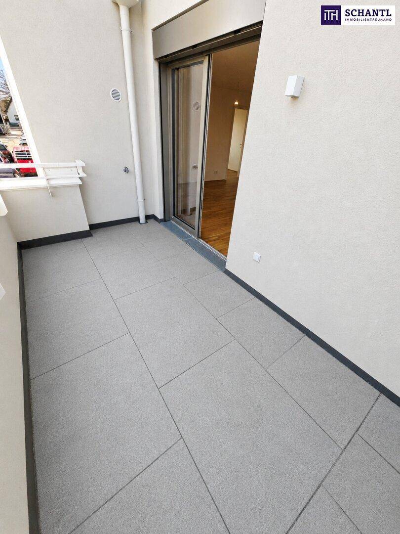 Geräumiger Balkon mit grauen Fliesen und Zugang zu einem Innenraum mit Holzfußboden durch eine Glasschiebetür.