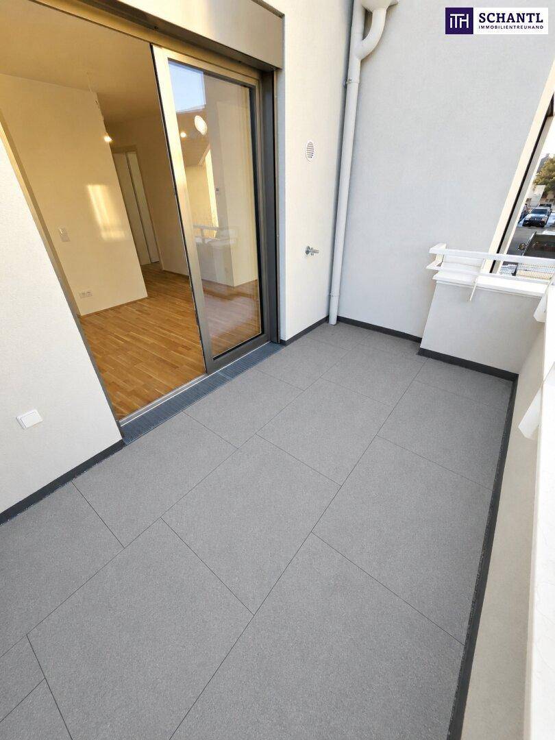 Heller Balkon mit grauen Fliesen und Glasschiebetür, die zu einem leeren Raum mit Holzfußboden führt.
