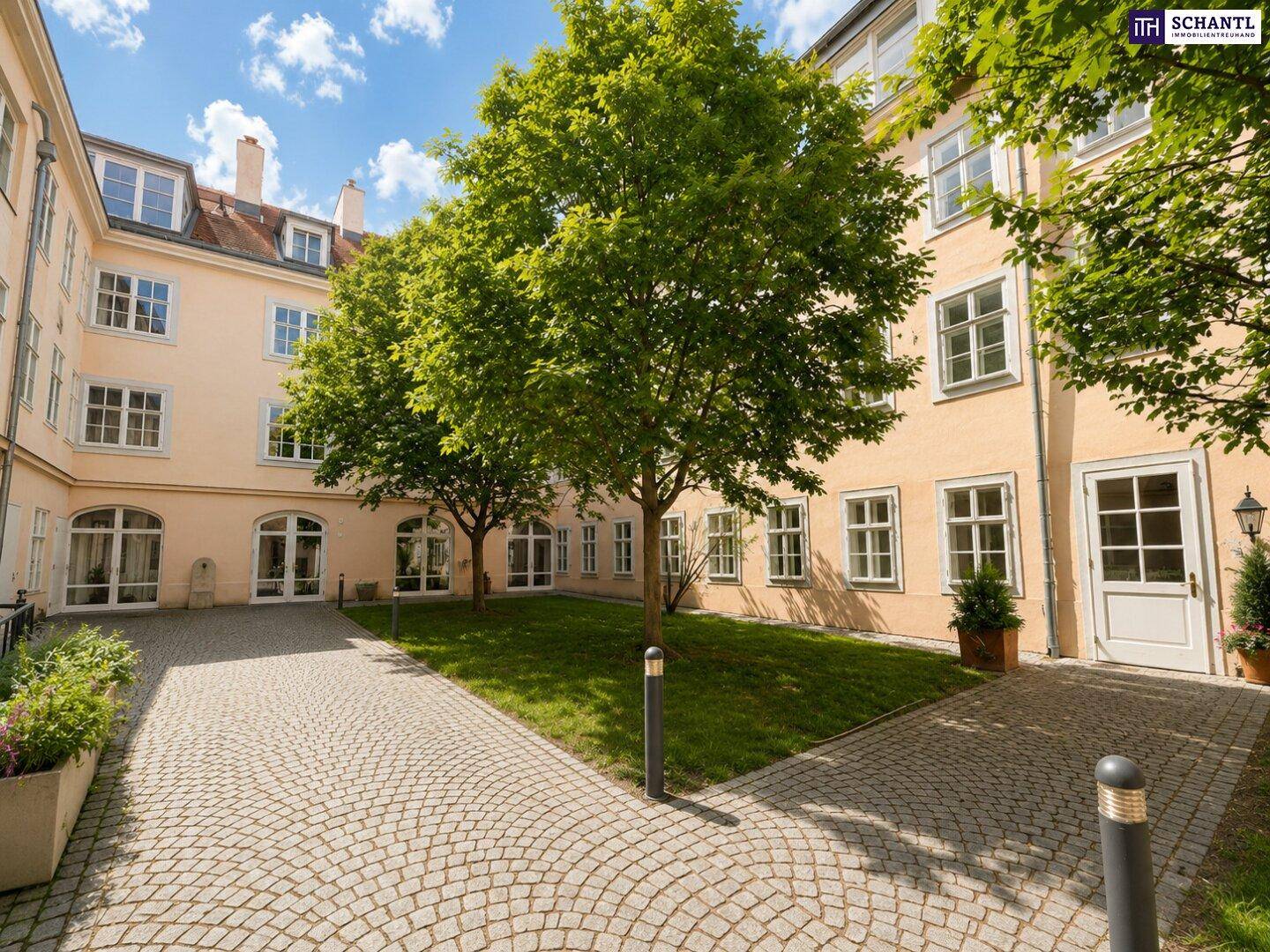 Idyllischer Innenhof mit Kopfsteinpflaster, grüner Rasenfläche und hohen Bäumen, umgeben von historischen Gebäudefassaden.