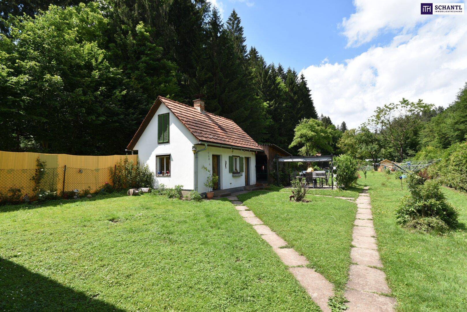 Ansicht des kleinen weißen Hauses von vorne, umgeben von einem gepflegten Garten und Wald.