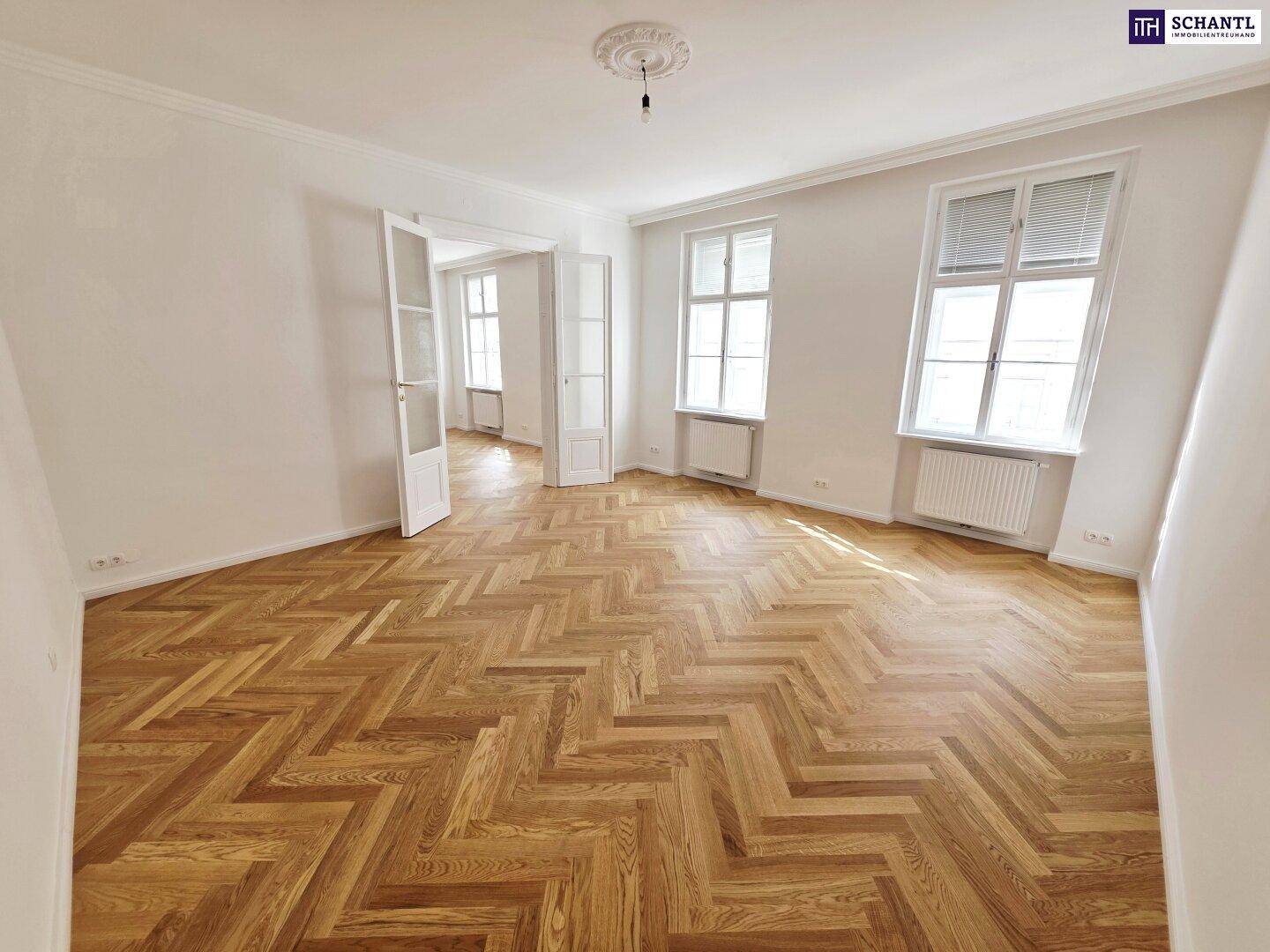 Lichtdurchflutetes Zimmer mit zwei großen Fenstern, Fischgrätparkett und hohen Decken in einem Altbau.