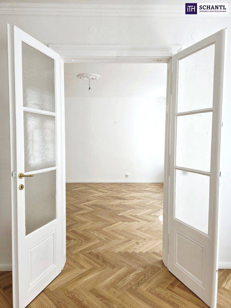 Blick durch eine Flügeltür in ein helles Zimmer mit Fischgrätparkett und Stuckdecke.