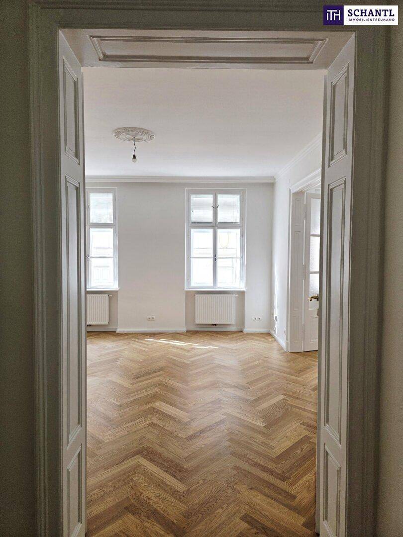 Blick durch eine Flügeltür in ein helles Zimmer mit Fischgrätparkett und zwei Fenstern.
