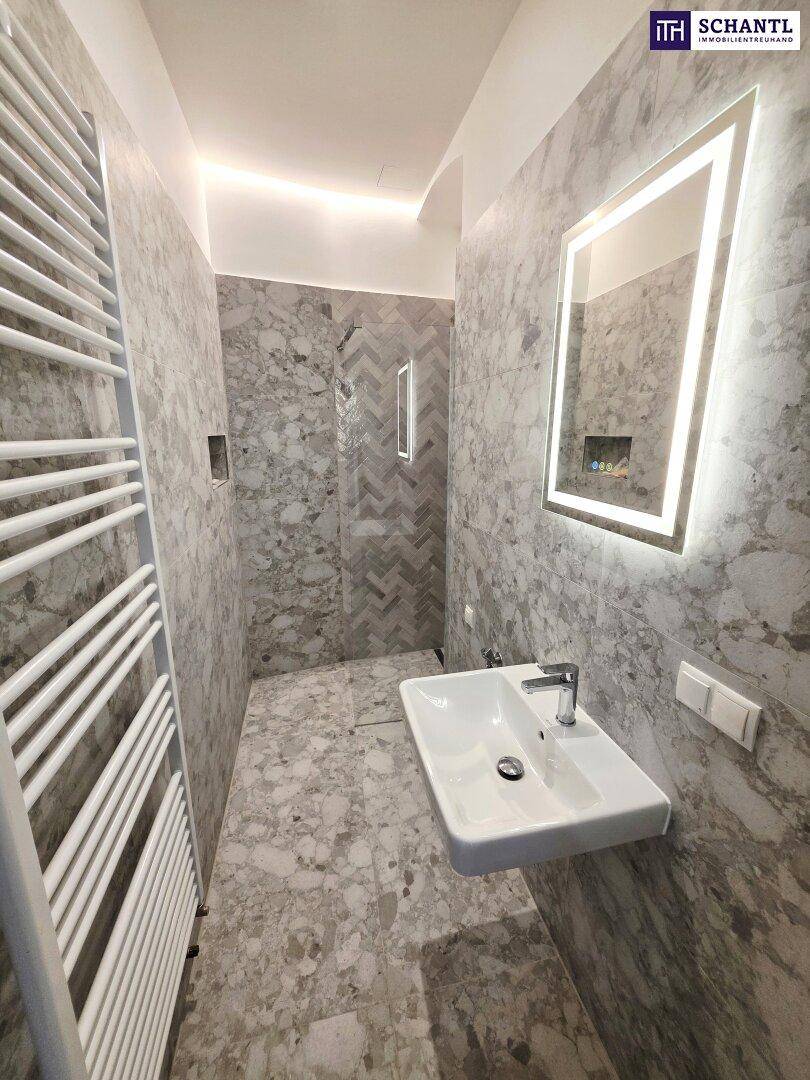 Modernes Badezimmer mit grauen Fliesen, beleuchtetem Spiegel und ebenerdiger Dusche.