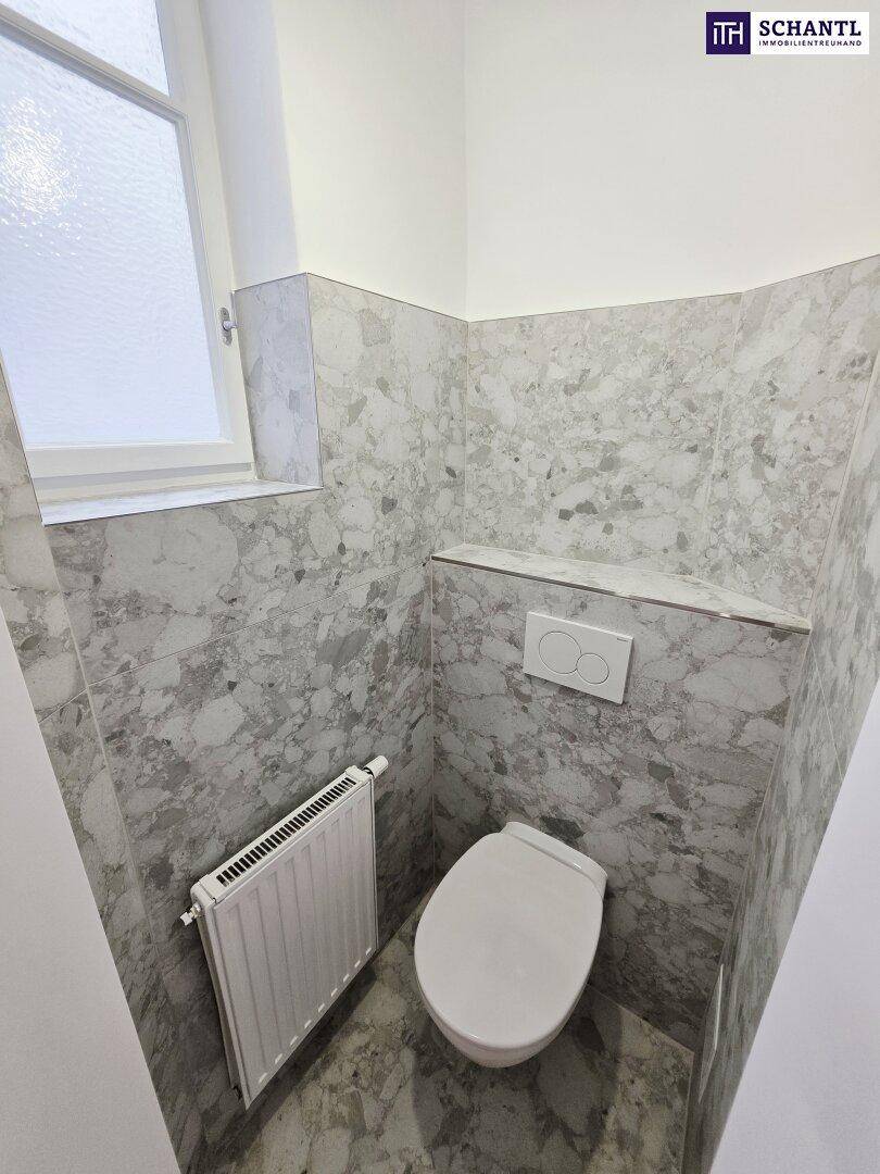 Modernes WC mit Wandtoilette, Fenster und grauen Fliesen, ideal als Gästetoilette.