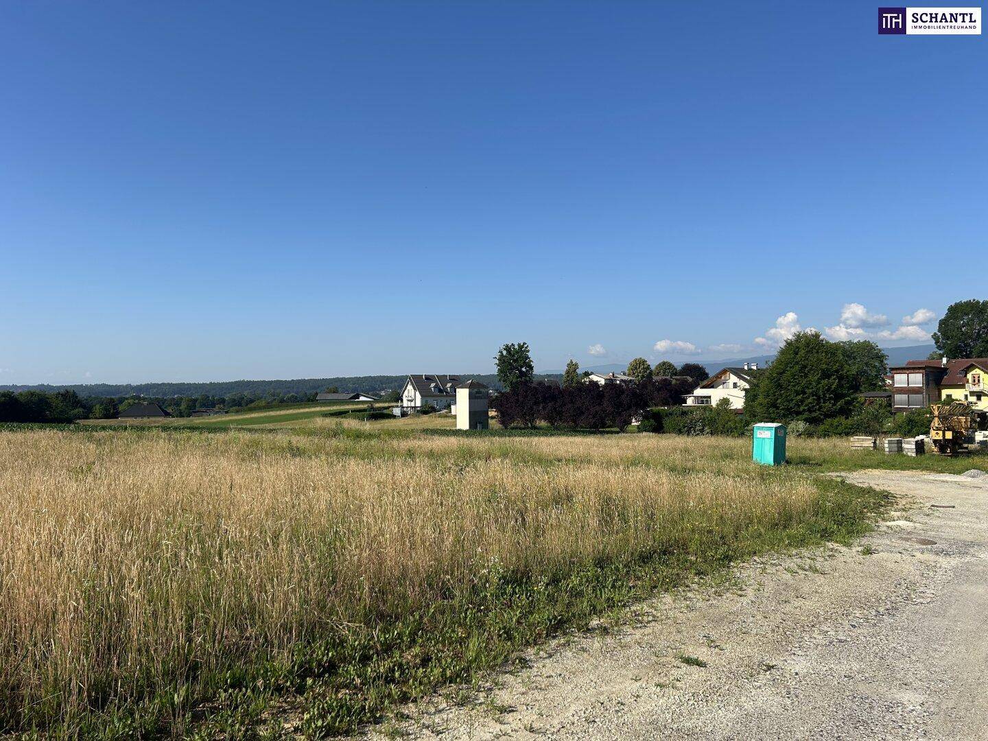 Grünfläche mit einem mobilen WC und Blick auf die umliegende Wohnbebauung und Landschaft.