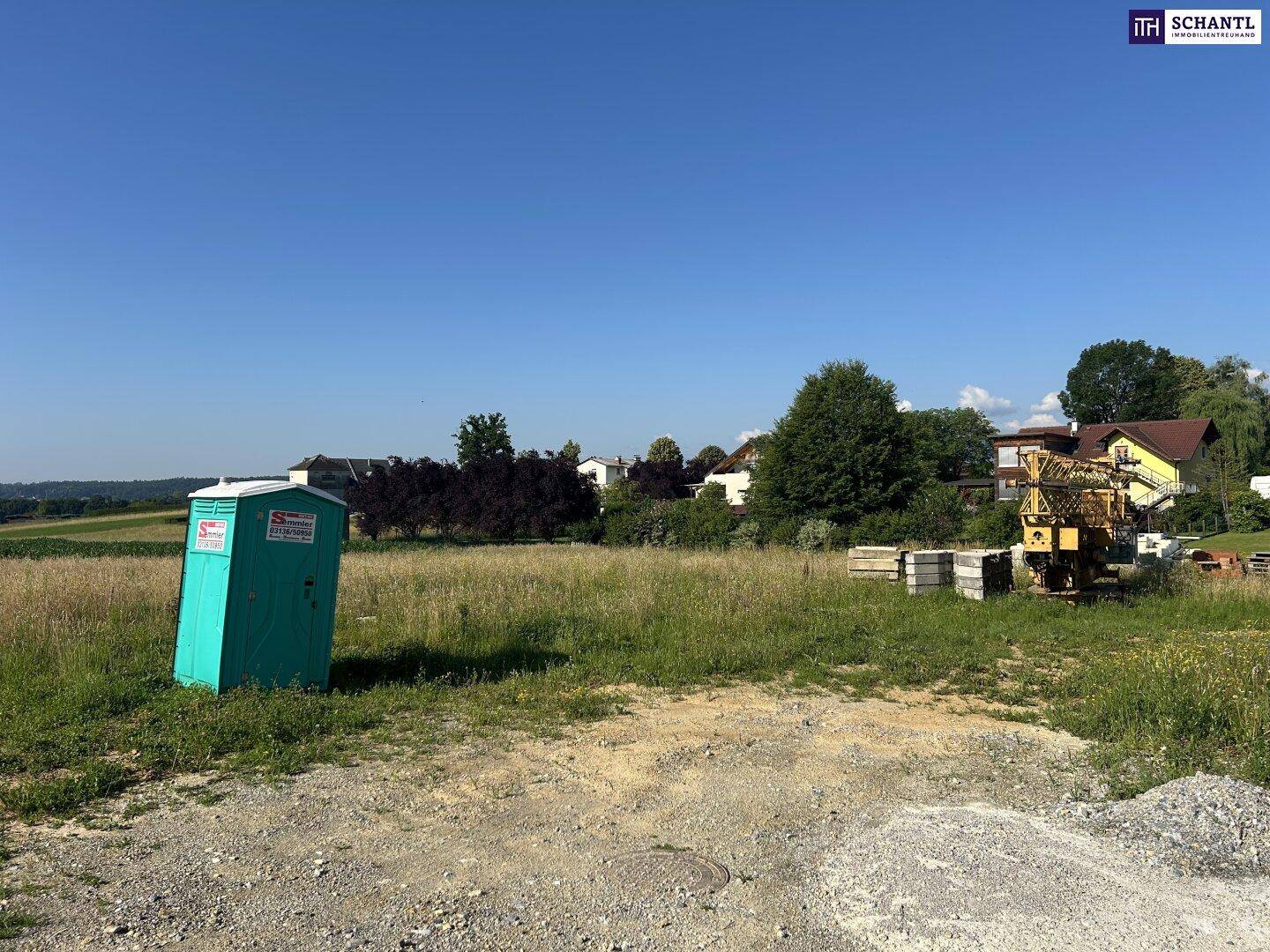 Feld mit mobilem WC, Baustoffen und einem Kran, umgeben von Bäumen und Häusern im Hintergrund.