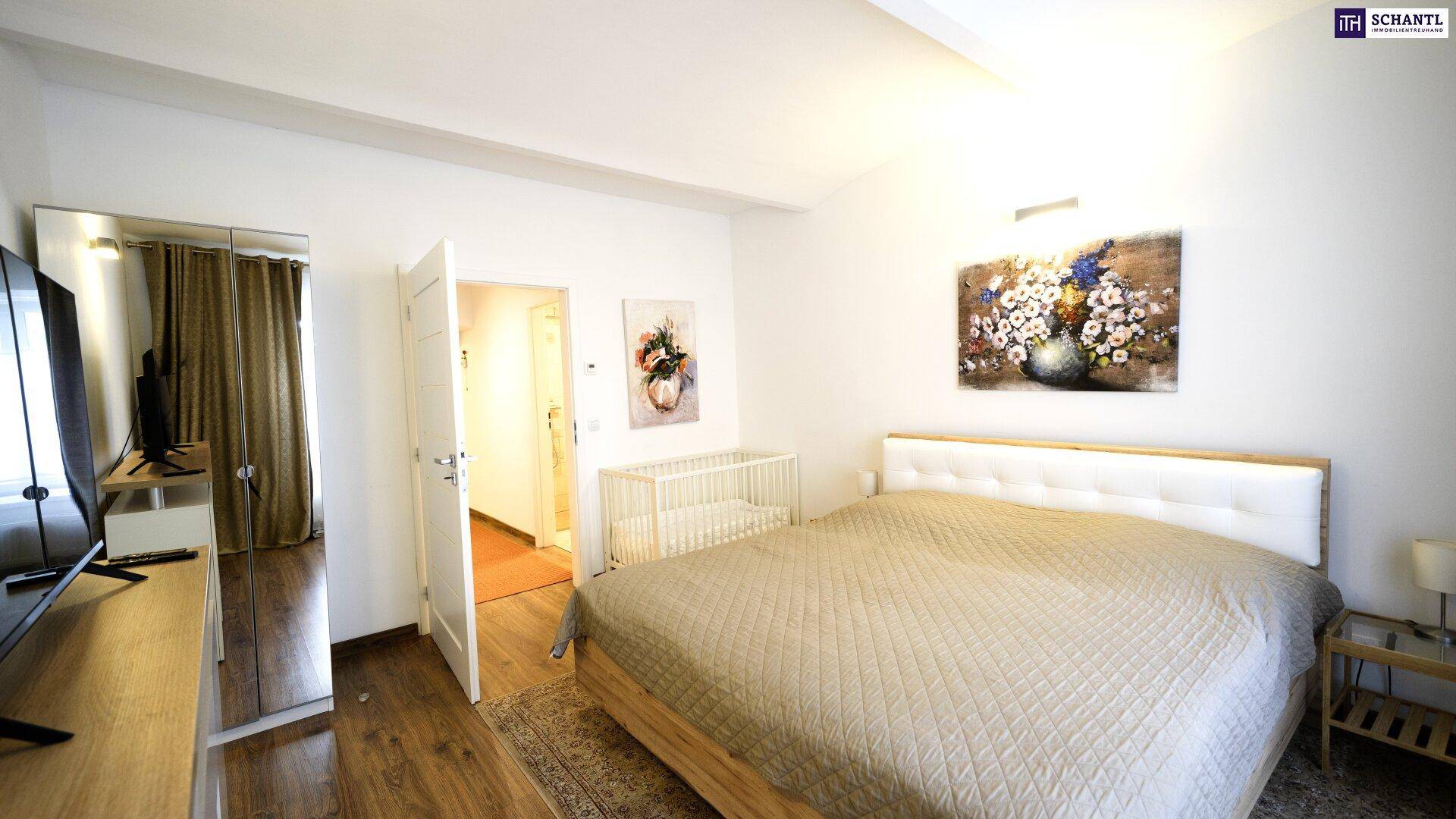 Helles Schlafzimmer mit Doppelbett, Holzboden und Blick auf ein Kinderbett im Nebenzimmer.