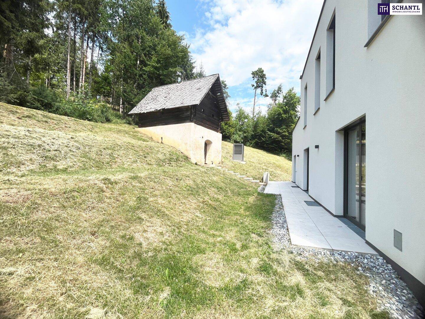 Moderne weiße Gebäudefassade neben einem grasbewachsenen Hang mit altem Holzschuppen und Bäumen.