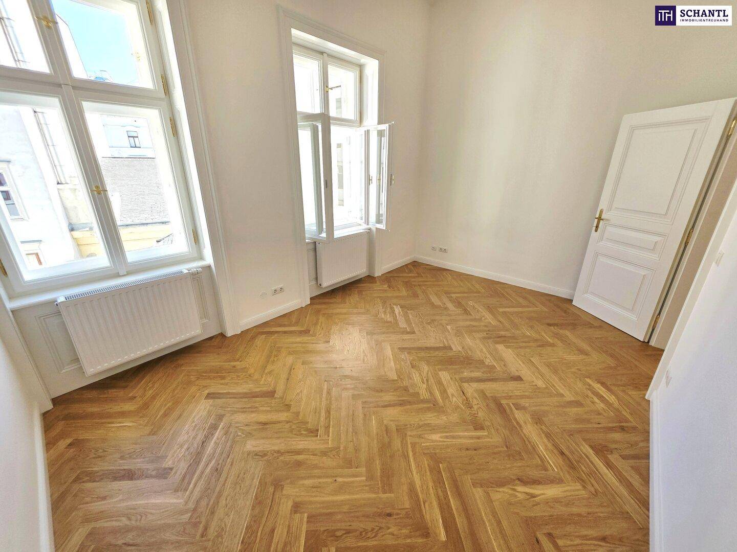 Helles Zimmer mit Fischgrätparkett und hohen Fenstern, das viel Tageslicht hereinlässt.