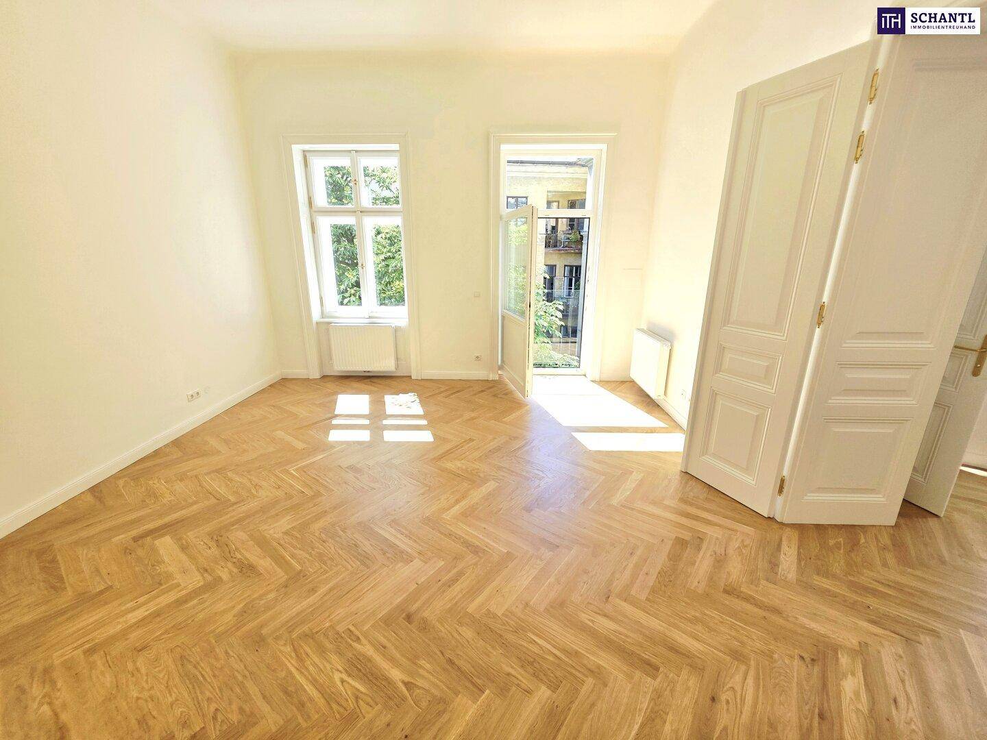 Helles Zimmer mit Fischgrätparkett und hohen Fenstern, das viel Tageslicht hereinlässt.